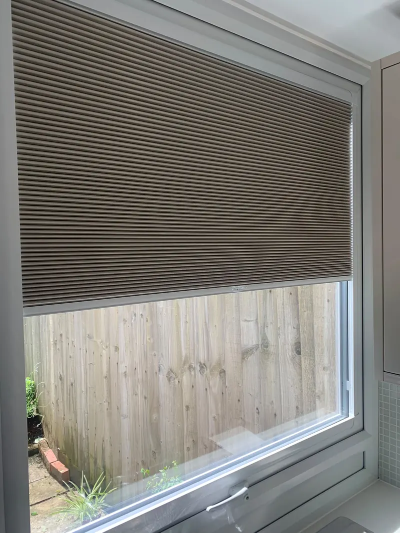 A pleated blind fitted to a kitchen window, part lowered above the garden fence
